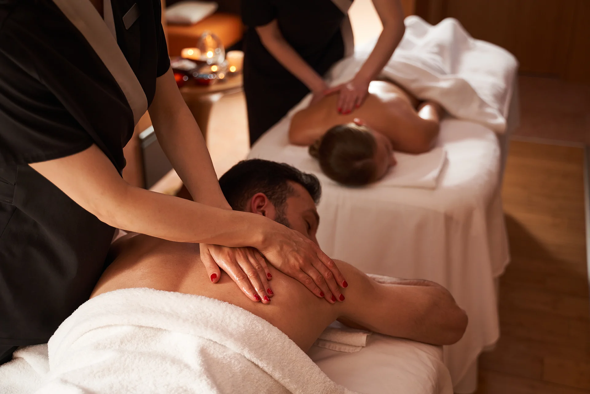 Over-the-shoulder view of a couple receiving full-body Swedish massages from two therapists in black uniforms. A man is in the foreground.